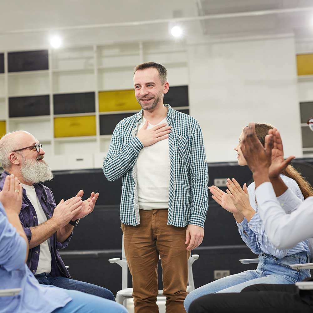 A member of a group therapy session stands up and talks while others clap for him